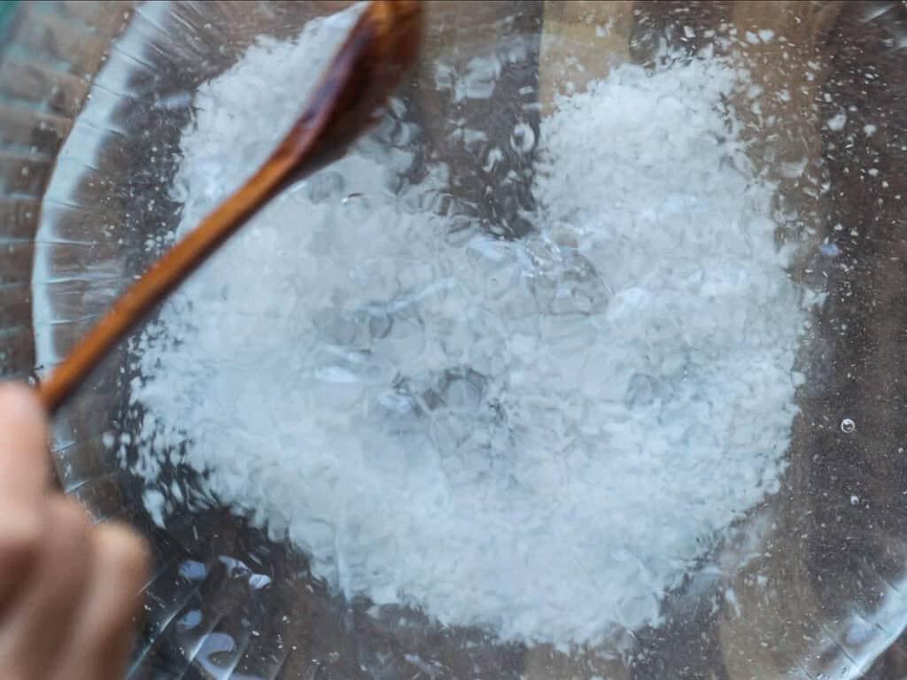 Close-up of a glass bowl with a white substance being mixed by a wooden spoon. The texture is granular and resembles salt surrounded by clear liquid. The bowl is on a wooden surface.